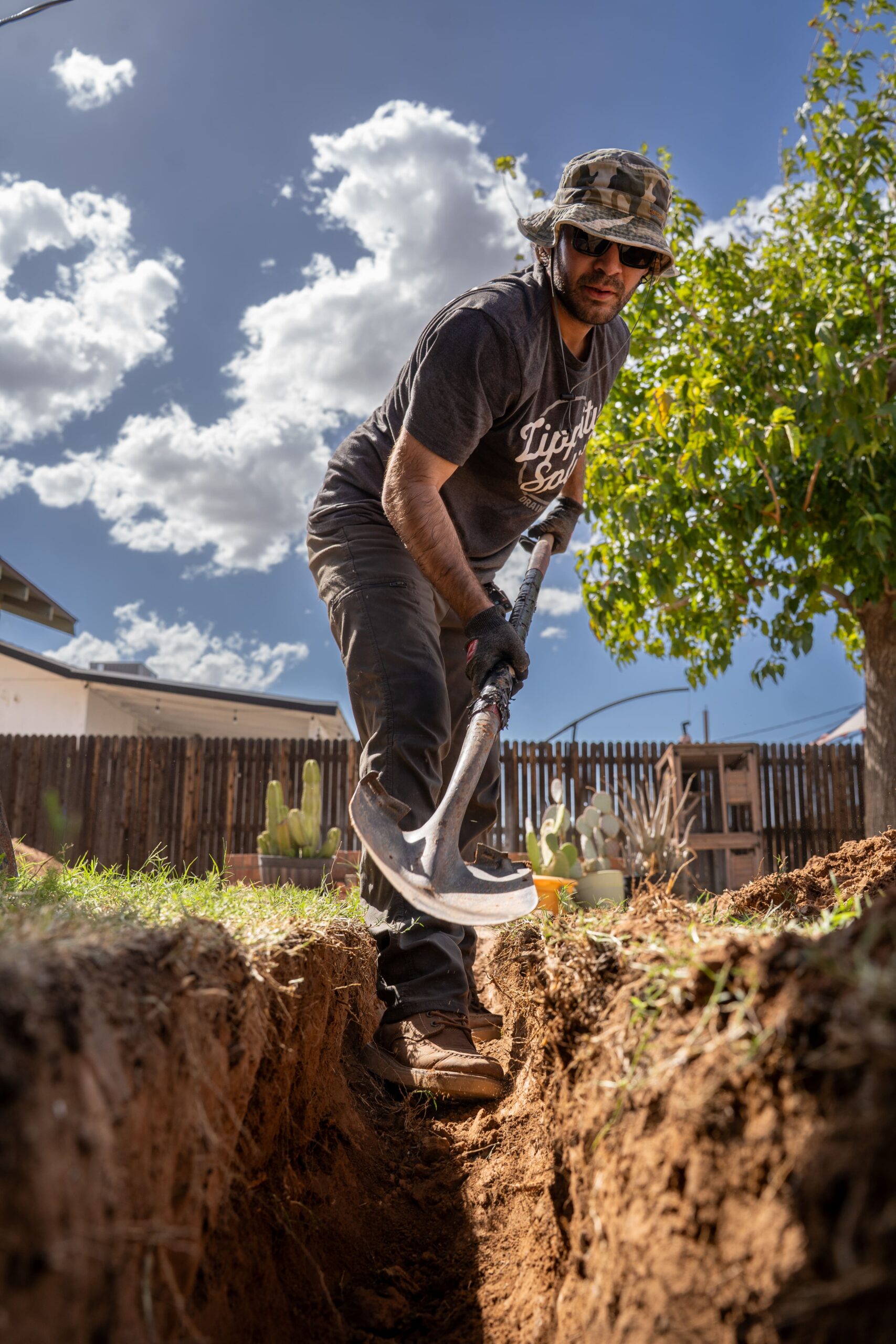 Man wearing a camo hat and sunglasses digging a deep trench with a shovel in a sunny backyard.